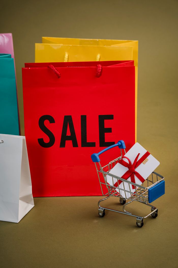 Colorful shopping bags with a mini cart holding a gift symbolize holiday sales and gift shopping.