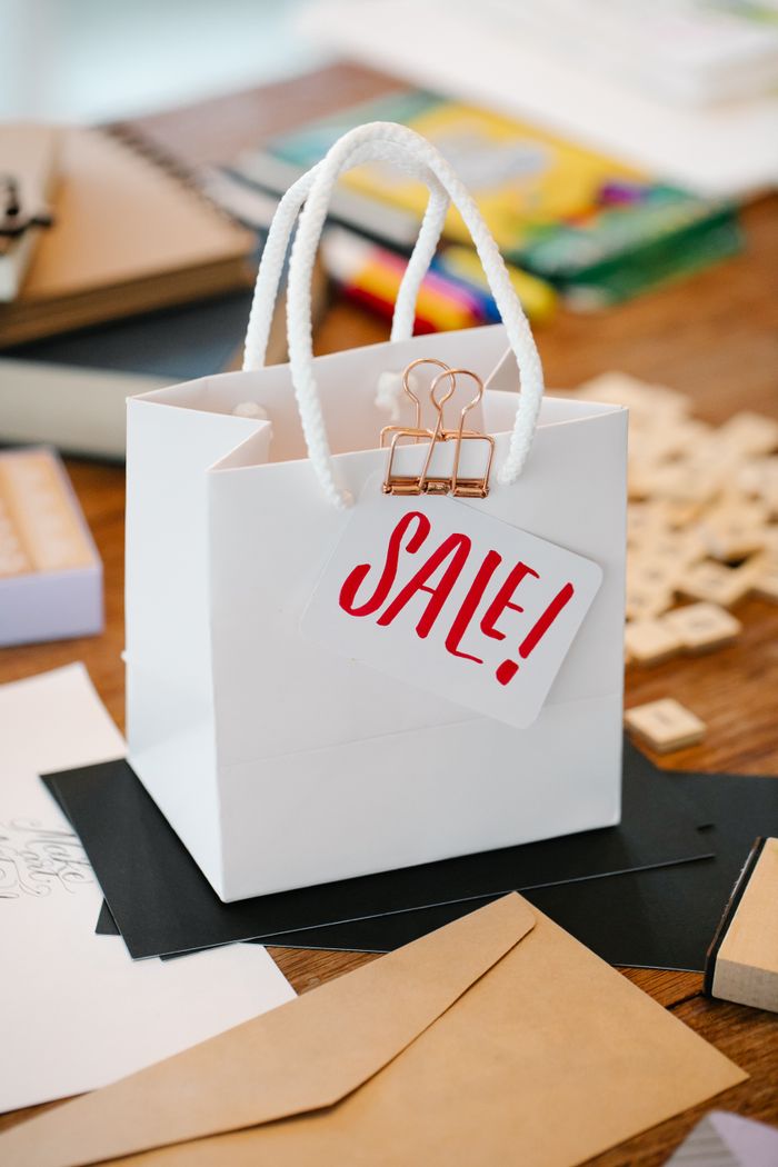 A white paper bag featuring a 'Sale!' tag on a wooden desk with stationery items.