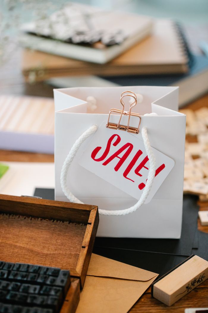 White paper bag with a red 'Sale!' tag on a wooden table surrounded by crafting materials.